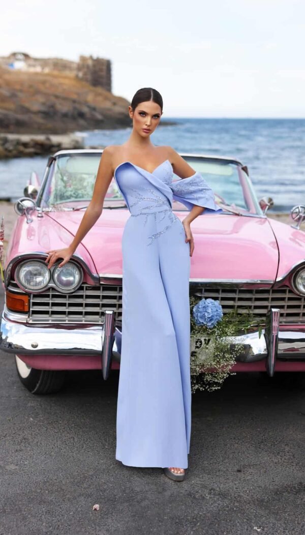 Model wearing a powder blue one-shoulder jumpsuit with wide-leg silhouette, standing by a vintage pink car – Casa Reina couture.