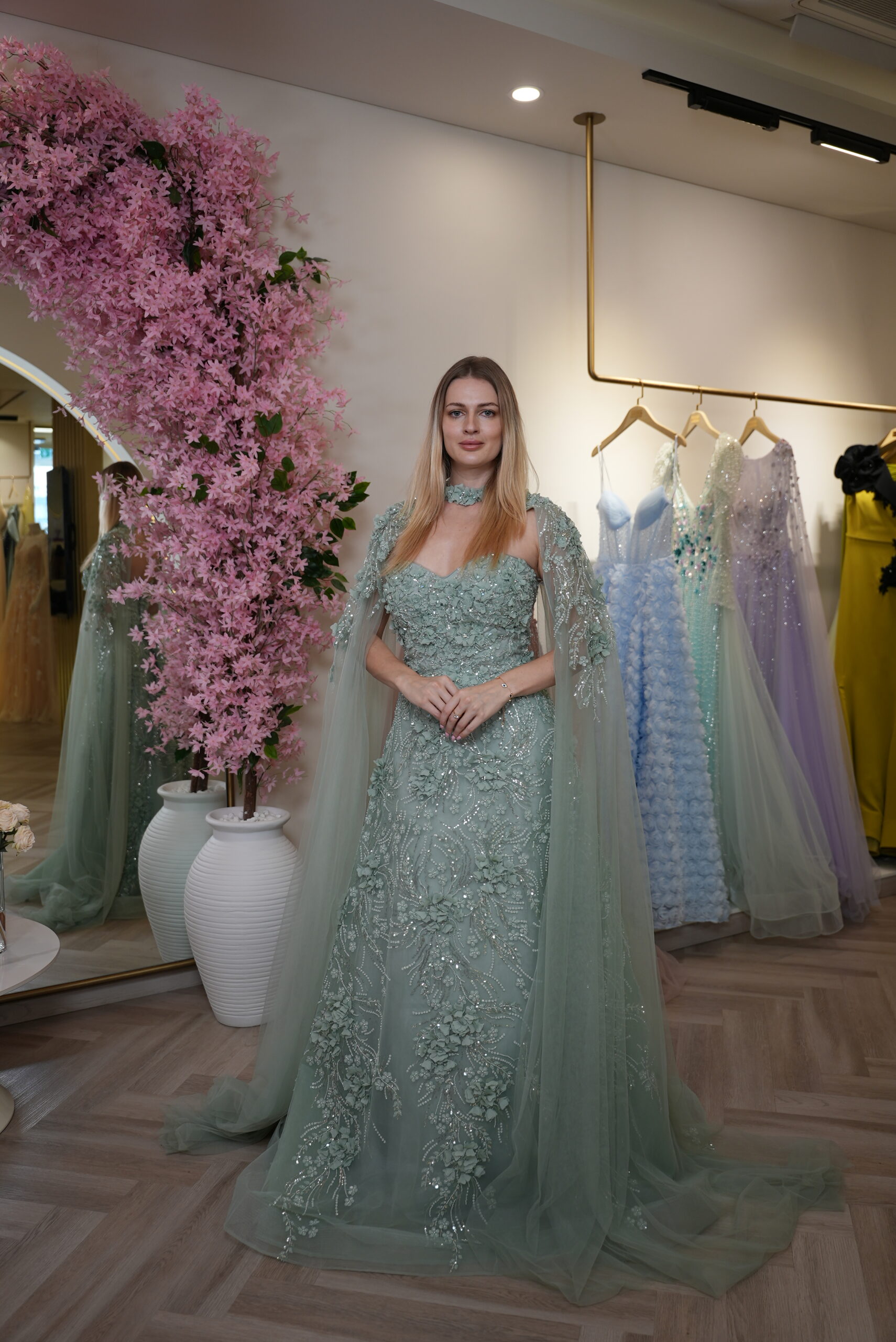 Model wearing an embroidered evening gown with 3D floral appliqué, illusion sweetheart neckline, and sheer cape sleeves in Sage Green – Casa Reina Boutique,