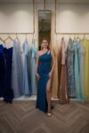 Model wearing a teal one-shoulder evening gown with cutout detail, ruched bodice, shimmering fabric, and thigh-high slit – Casa Reina Boutique