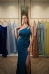 Model wearing a teal one-shoulder evening gown with cutout detail, ruched bodice, shimmering fabric, and thigh-high slit – Casa Reina Boutique