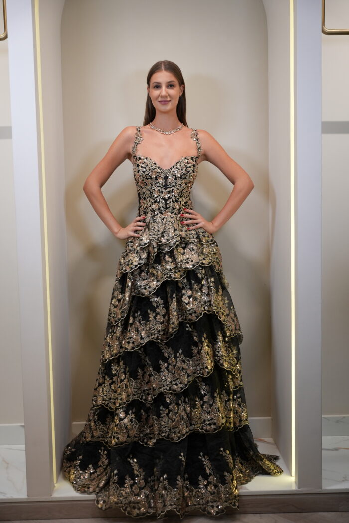 Model wearing a black and gold tiered floral embroidered evening gown with sweetheart neckline and layered skirt – Casa Reina couture fashion.