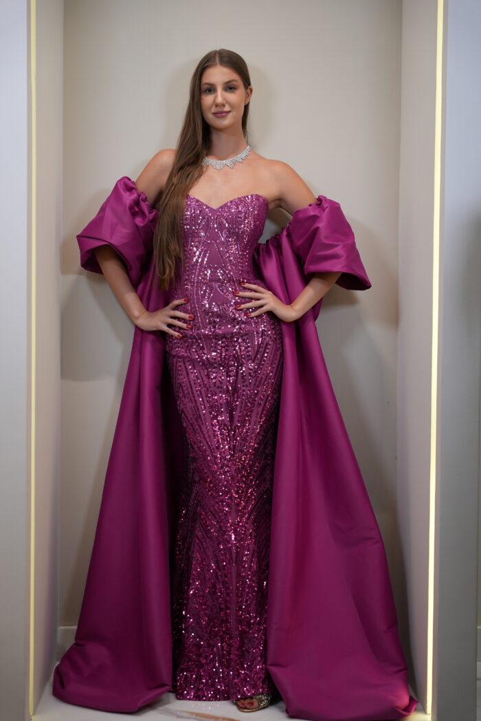 Model wearing a magenta strapless sequin evening gown with sweetheart neckline, detachable puff sleeves, and flowing cape – Casa Reina couture collection.