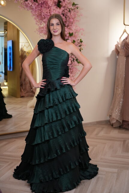 Model wearing an emerald green one-shoulder evening gown with floral applique detail and cascading tiered ruffle skirt – Casa Reina couture collection.