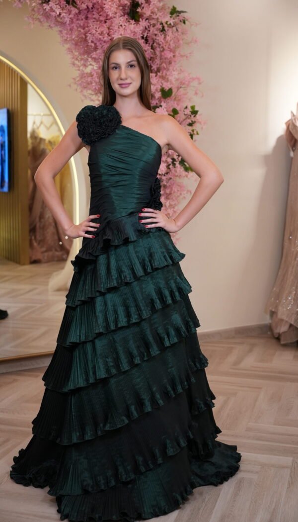 Model wearing an emerald green one-shoulder evening gown with floral applique detail and cascading tiered ruffle skirt – Casa Reina couture collection.