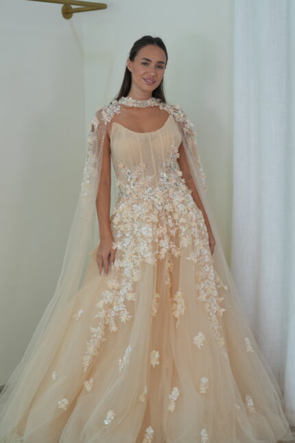 Model wearing a blush evening gown with pleated bodice, floral appliqués, crystal embellishments, and sheer embellished cape – Casa Reina Boutique