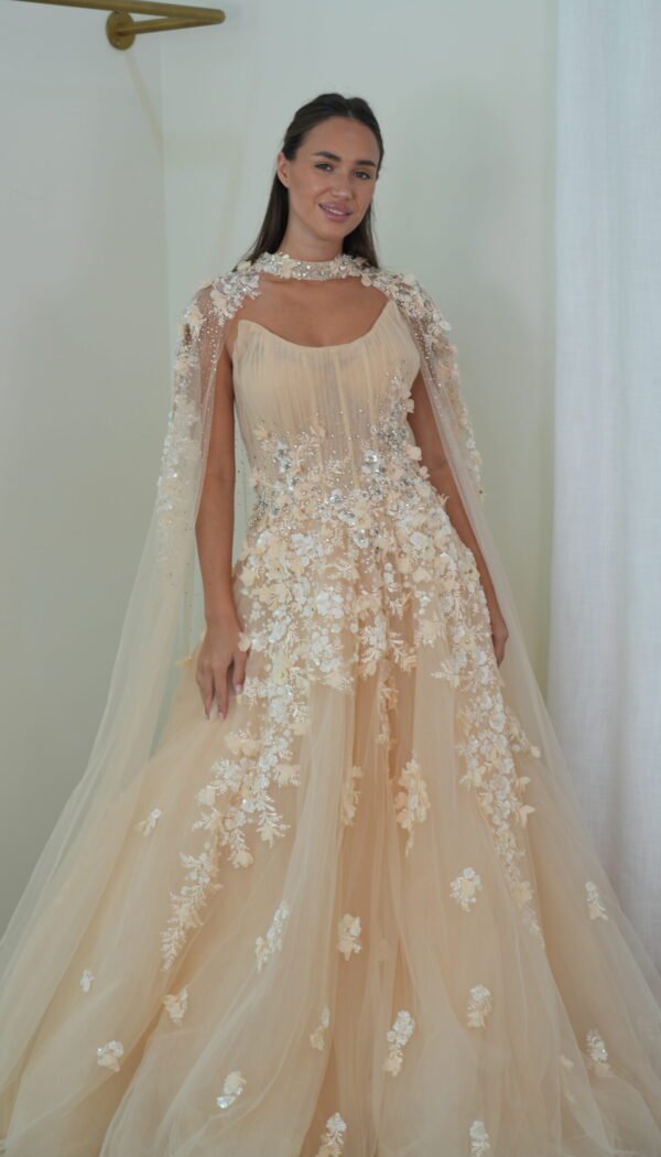 Model wearing a blush evening gown with pleated bodice, floral appliqués, crystal embellishments, and sheer embellished cape – Casa Reina Boutique