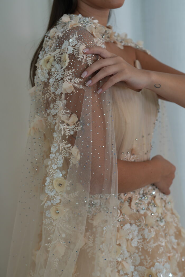 Model wearing a blush evening gown with pleated bodice, floral appliqués, crystal embellishments, and sheer embellished cape – Casa Reina Boutique