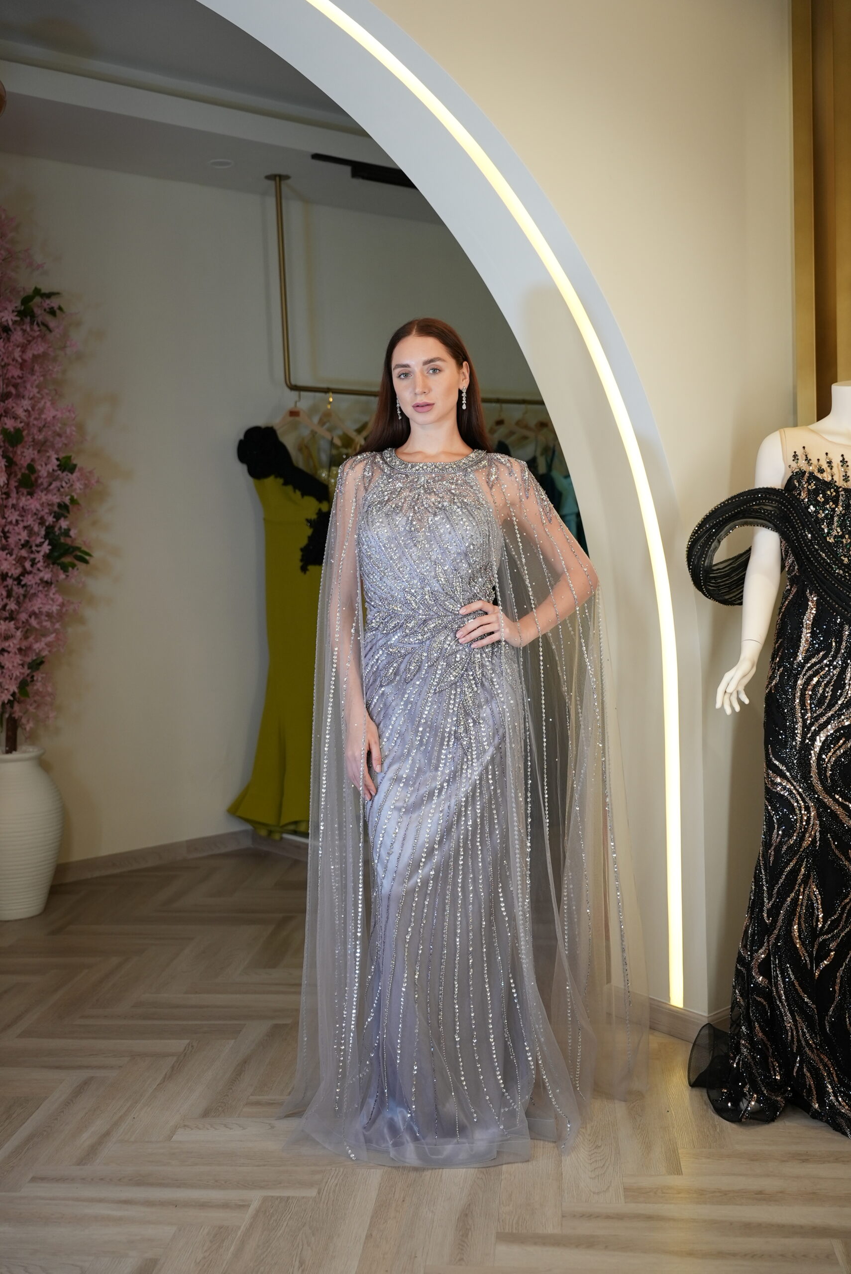 Model wearing a silver embellished evening gown with sheer beaded cape overlay and fitted silhouette – Casa Reina couture collection.