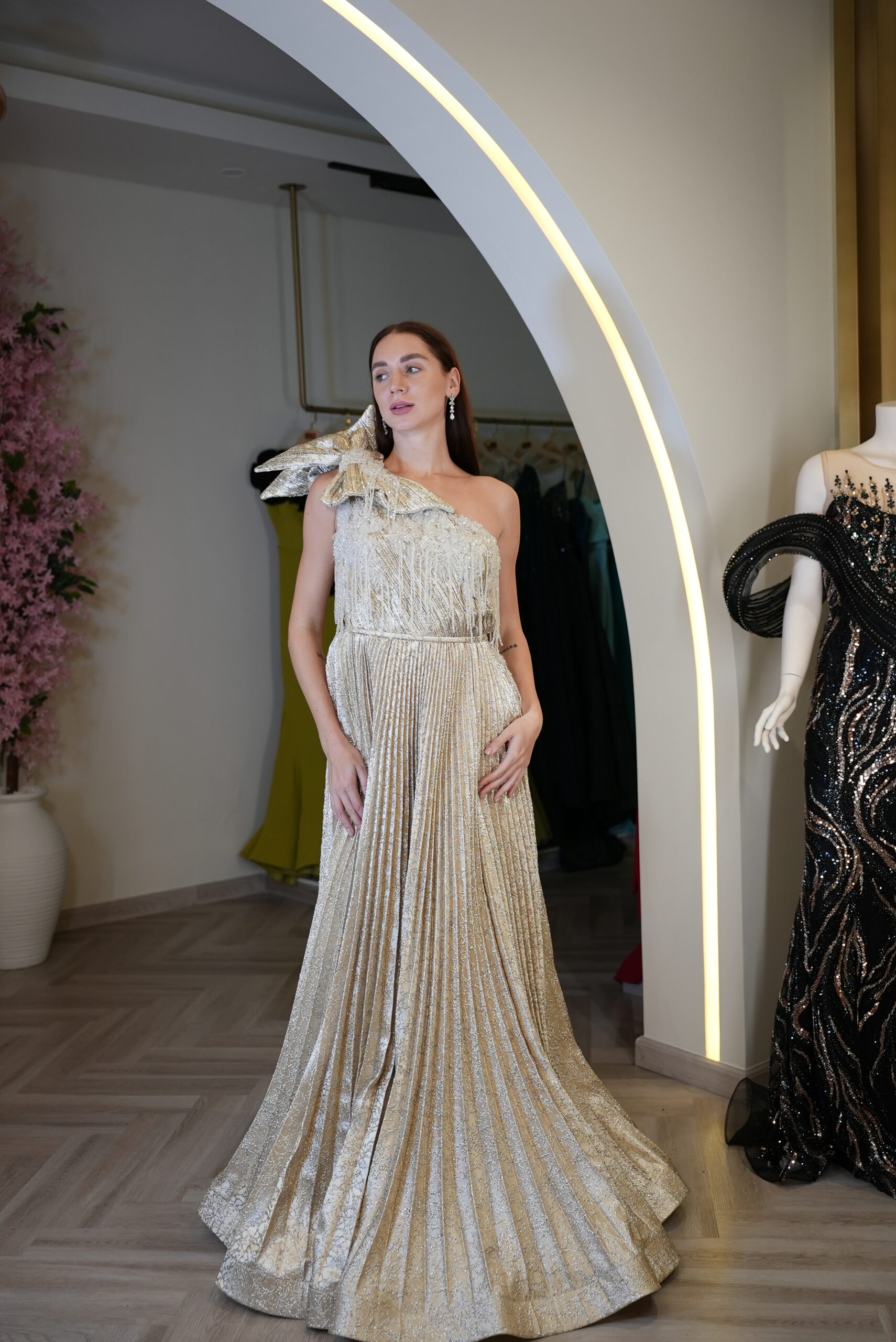 Model wearing a metallic gold one-shoulder evening gown with fringe bodice, pleated full skirt, and bow accent – Casa Reina Boutique