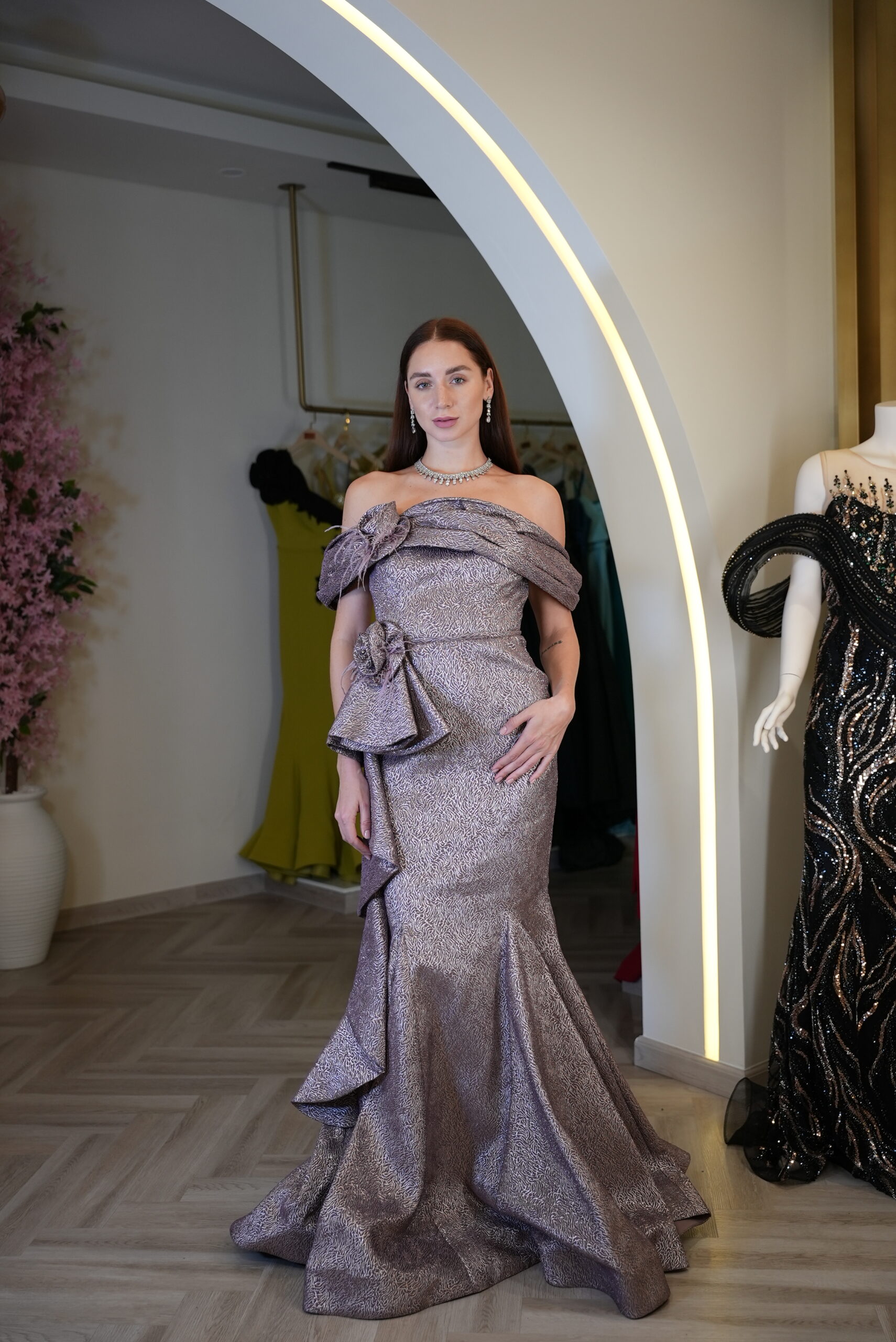 Model wearing a metallic mauve off-shoulder evening gown with layered ruffles, floral rosettes, and fitted mermaid silhouette – Casa Reina Boutique