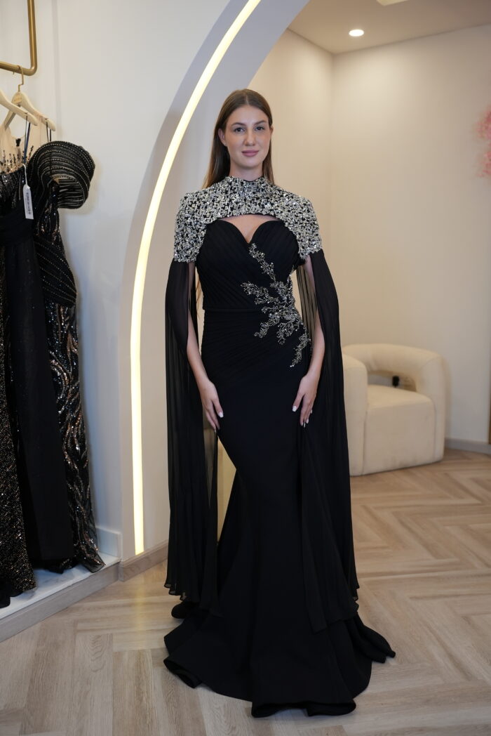 Model wearing a black evening gown with a sweetheart neckline, silver embellished bodice, and flowing cape sleeves