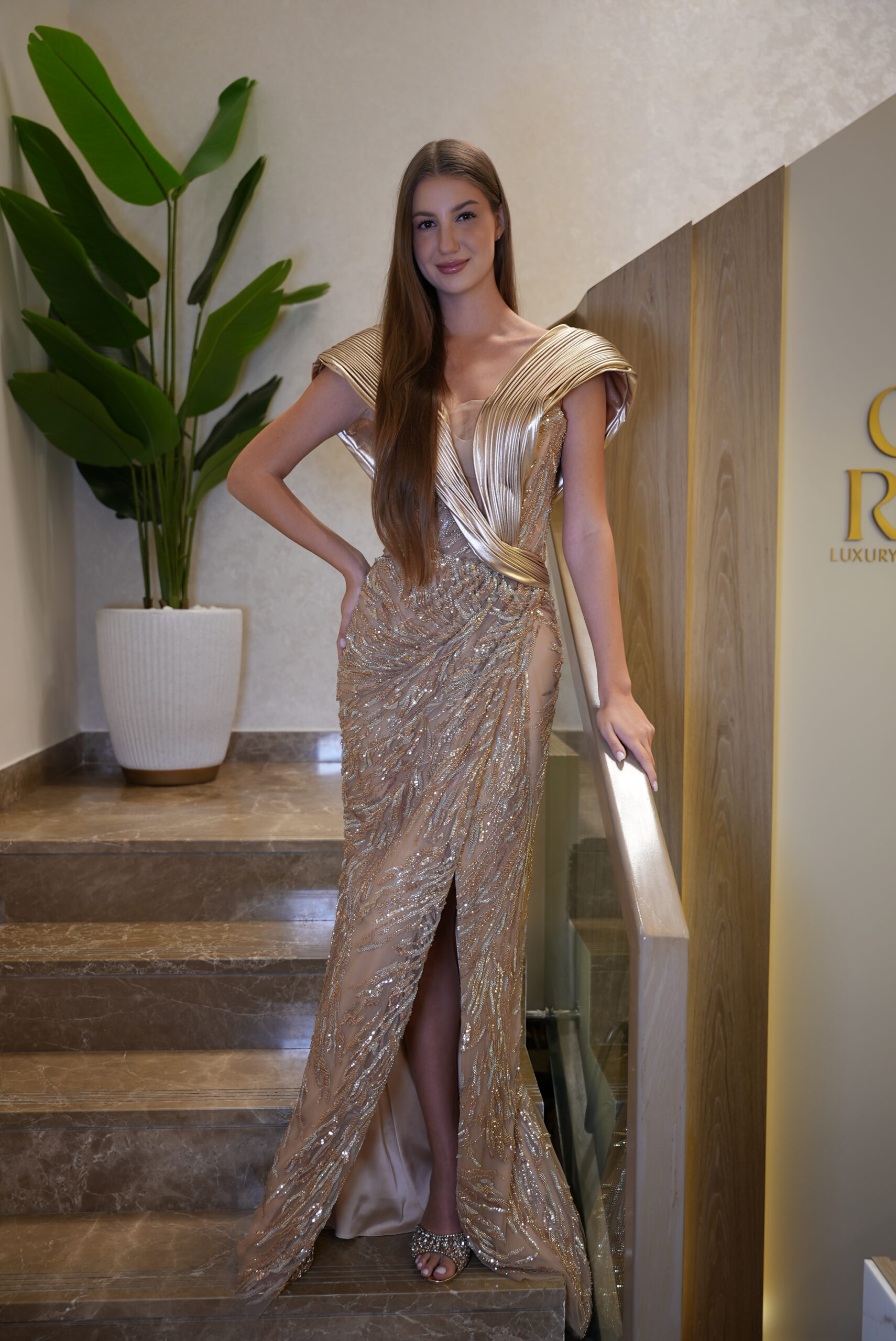 Model wearing a golden metallic pleated evening gown with sculpted shoulders and embellished sequin skirt featuring a thigh-high slit